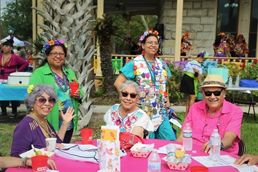 A group of people sitting at a table, with two ladies and a man seated, and two other ladies standing beside them at a fiesta.