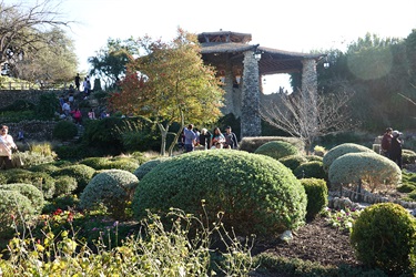 A diverse group of people standing around a lush garden next to the stone gazebo, chatting and enjoying the beautiful scenery at the Japanese Tea Garden.