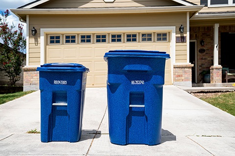 Two blue trash cans, one larger than the other, are seen in front of a house.