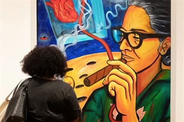 A person stands in front of a large painting of a woman with glasses.